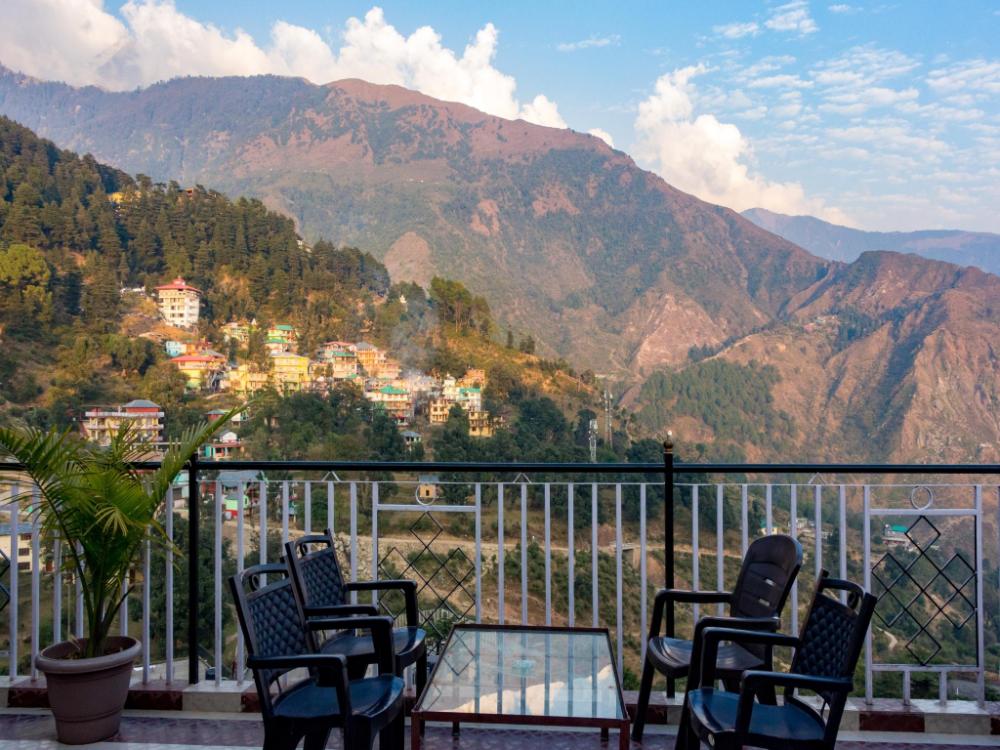 Private balcony with two plastic chairs and a table overlooking the Dhauladhar range and village